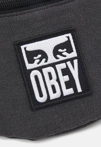 Μαύρο ύφασμα με μια λευκή κεντητή απεικόνιση δύο χεριών και τη λέξη "OBEY" με κεφαλαία γράμματα στο κέντρο.