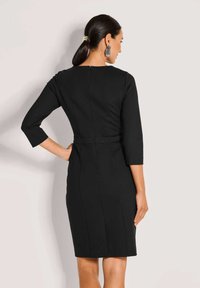 Zwarte jurk met driekwart mouwen, nauwsluitend ontwerp, knielengte, voorzien van verticale naden en een rits aan de achterzijde in het midden. Gladde stoftextuur.