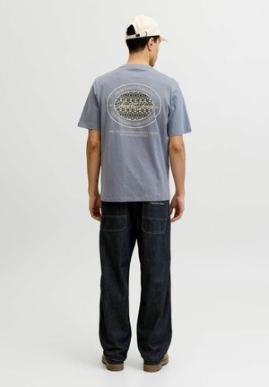 Uomo che indossa una t-shirt grafica viola chiaro, jeans scuri e larghi, stivali color cuoio chiaro e cappellino da baseball bianco, in piedi con le spalle rivolte a uno sfondo bianco.