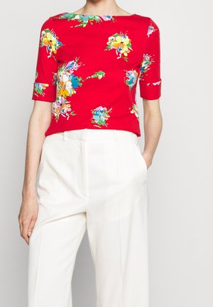 Haut rouge à manches courtes avec imprimés floraux colorés, associé à un pantalon blanc taille haute, une main dans la poche.