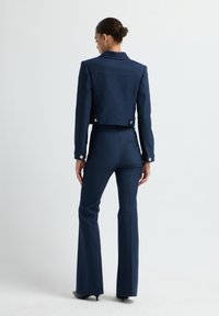 Žena s tmavými vlasy v drdolu, oblečená v přiléhavém námořnickém blejzru a odpovídajících rozšiřujících se kalhotách, stojící zády k fotoaparátu.