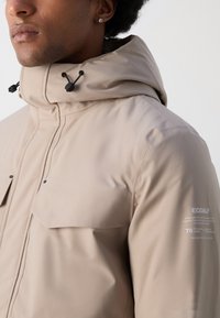 Chaqueta impermeable beige con alto cuello, capucha ajustable con cordón, dos bolsillos delanteros y una marca Ecoalf en la manga.