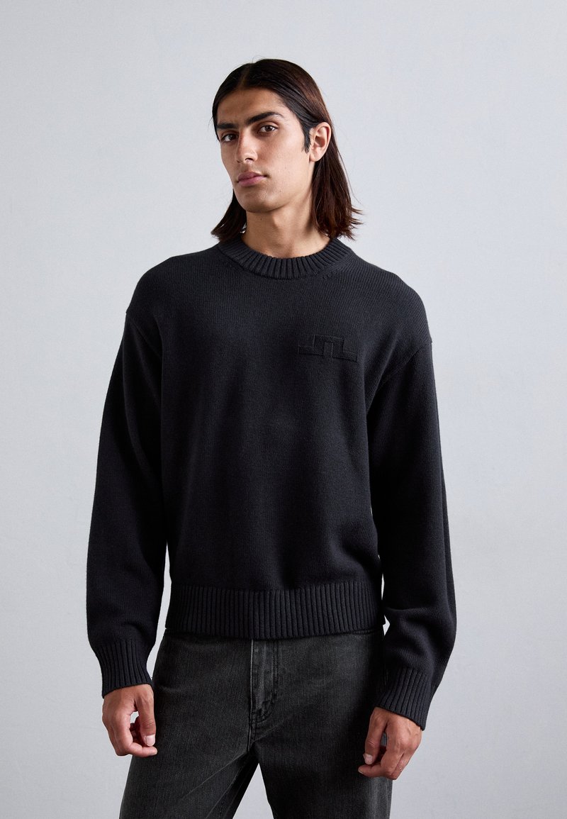 Black knit sweater with a ribbed neckline and hem, featuring a subtle logo patch on the chest and wide cuffs. Paired with dark jeans.