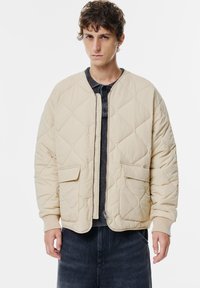 Veste en tweed beige matelassée en tissu texturé, dotée d'une fermeture éclair à l'avant, de deux poches avant et de poignets côtelés, portée sur une chemise sombre.