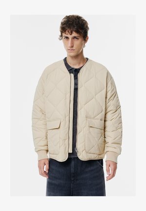 Beige vuorattu takki, joka on valmistettu teksturoidusta kankaasta, jossa on vetoketjukiinnitys, kaksi etutaskua ja ribbireunat, puettuna tumman paidan päällä.