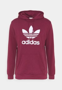 Mörkröd hoodie med framficka, justerbar dragsnodd i huvan och vit Adidas-logotyp med tre ränder och en trefoil-design.