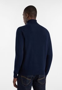 Temno modra pulover s visokim ovratnikom, izdelan iz mehke tkanine. Ima dolge rokave in rebraste manšete. Obdan je s temnimi kavbojkami.