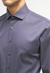 Camicia uomo di colore viola scuro con bottoni, realizzata in tessuto morbido, caratterizzata da un colletto classico e un logo CK ricamato sul petto.