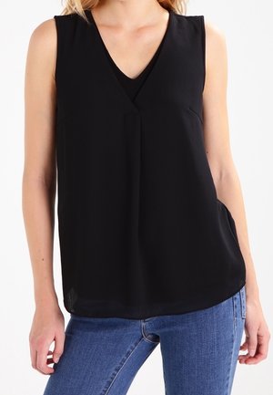 Mouwloze zwarte V-hals blouse gedragen met blauwe spijkerbroek, met zichtbaar bovenlichaam en armen tegen een effen lichte achtergrond.