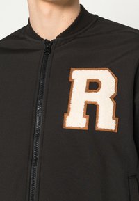 Svart bombarjacka med en texturerad, krämfärgad "R"-patch med bruna accenter och en framdragbar dragkedja. Mjukt material och figurnära design.