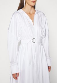 Hvid, langærmet kjole med krave, indsnøring i taljen og bælte med en sølvfarvet rund spænde; glat bomuldsstof med plisserede detaljer.