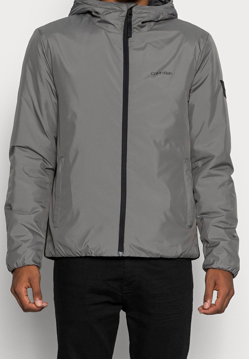 Homme portant une veste à capuche grise Calvin Klein avec une fermeture éclair frontale et des poches latérales, associée à un pantalon noir.