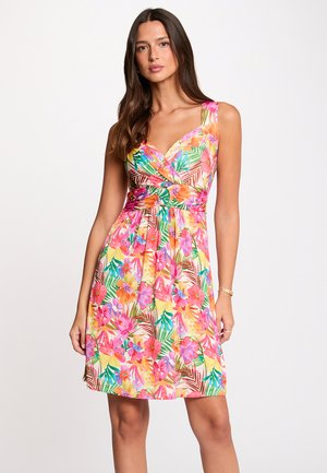 Donna che indossa un vestito senza maniche, lungo fino al ginocchio, con un vivace motivo floreale e foglie rosa, arancioni e verdi, in piedi su uno sfondo bianco uniforme.