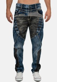 Pantalons cargo en denim avec un délavage bleu, présentant des panneaux en camouflage, des coutures décoratives, des boutons latéraux et des accents noirs contrastants.