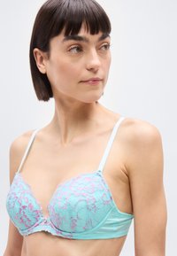 Γυναίκα με κοντά σκούρα μαλλιά που φοράει ανοιχτό μπλε δαντελένιο σουτιέν με ροζ λουλουδάτες λεπτομέρειες, κοιτάζοντας ελαφρώς προς το πλάι, με φόντο απλό.