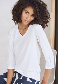 Weißes V-Ausschnitt-Shirt mit Dreiviertelärmeln, weicher Textur, dezente Markenkennzeichnung am Ärmel, kombiniert mit gemusterten blauen Hosen.