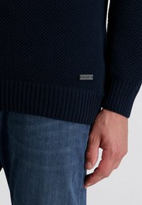 Navy blauwe gebreide trui met een gestructureerde afwerking, voorzien van een geribbelde onderkant en manchetten. Klein label rechtsonder met de merknaam.