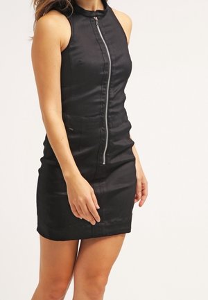 Robe mini noire sans manches avec une fermeture éclair à l'avant, taille cintrée et poches dissimulées, portée par une personne debout sur un fond uni.