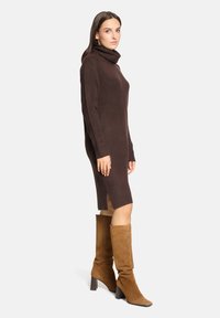 Femme debout en robe-pull marron à col roulé et bottes hautes en daim beige, regardant légèrement sur le côté devant un fond uni.
