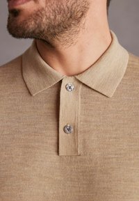 Beige polo-paita, jossa on kaulus, kolmesta napista koostuva kaula-aukko ja teksturoitu kangaspinta. Hopeiset napit korostavat muotoilua.