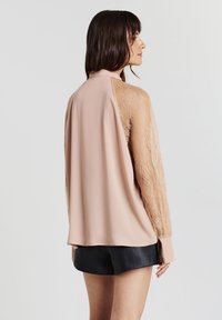 Frau seitlich stehend, mit einer beigen Bluse mit Spitzenärmeln und schwarzen Shorts vor einem schlichten hellen Hintergrund.