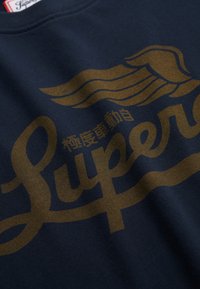 Marineblauw textiel met een goudkleurige gestileerde vleugelafbeelding en grote tekst "Super" naast kleine Japanse karakters.