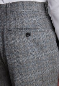 Gray plaid trousers featuring a textured fabric. Includes a rear pocket with a black button detailing. Subtle brown accents in the pattern.