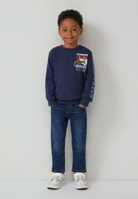 Felpa navy con stampa grafica e testo sulla parte anteriore, abbinata a jeans blu e sneakers bianche con accenti grigi. Texture liscia.