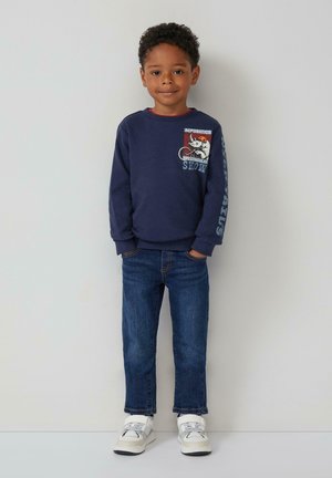 Marineblauwe sweatshirt met grafische print en tekst op de voorkant, gecombineerd met blauwe jeans en witte sneakers met grijze accenten. Zachte textuur.