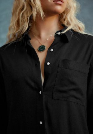 Camicia nera in lino con bottoni, dotata di taschino sul petto, caratterizzata da bottoni bianchi e una collana a strati con un pendente verde.