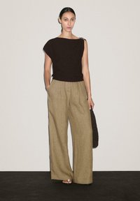 Haut asymétrique marron avec un accent sur l'épaule, associé à un pantalon large plissé beige. Le modèle est debout, les mains dans les poches, tenant un sac texturé.
