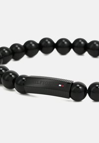 Bratara din mărgele negre, cu mărgele rotunde lucioase și o piesă de accent texturată de culoare neagră, înscripționată cu "HILFIGER" și culorile logo-ului.