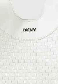 Hvit vevd håndveske med en glatt tekstur, buet topp og sølv DKNY-logo. Har et vevd mønster og strukturert design.