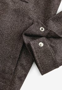 Bruine stof met fijne krijtstrepen en een gestructureerde afwerking. Voorzien van een knoopsluiting aan de manchette met glanzend metalen beslag.