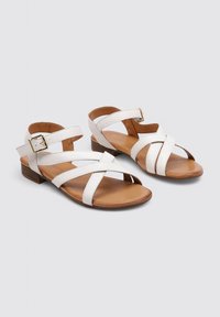 Sandales en cuir blanc avec des lanières entrecroisées et un petit talon. Les semelles intérieures sont brunes et il y a un détail de boucle métallique sur la lanière.