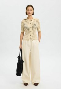 Cardigan beige en maille avec des manches mi-longues, devant à boutons et col ; associé à un pantalon large crème et un sac tote tissé noir.