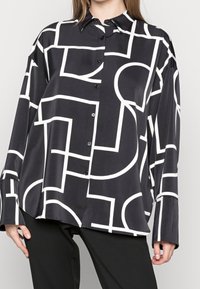 Blusa negra con patrones geométricos blancos, con un frente abotonado, mangas largas y una textura de tela suave y brillante.