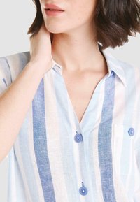 Camisa ligera a rayas en azul y blanco que cuenta con un frente abotonado, un bolsillo en el pecho y botones de tela. Material de lino texturizado.