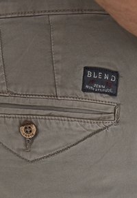 Beige denimshorts med en baklomme med en mørk stoffetikett som leser "B L E N D," akkompagnert av rød søm og en dekorativ knapp.