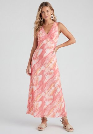 Frau in einem ärmellosen, rosa-weißen Batik-Maxi-Kleid mit Spitzenbesatz, stehend mit der Hand in der Hüfte, getragen große runde Ohrringe und Sandalen.