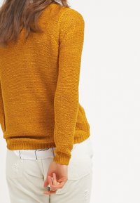 Maglione lavorato a maglia giallo senape con un motivo testurizzato, polsini aderenti e orlo dritto, abbinato a pantaloni bianchi con dettagli delle tasche visibili.