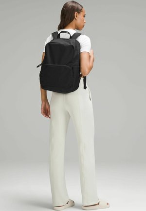 Čierny nylonový batoh s dvojitými bočnými vreckami, predným zipsovým oddelením, nastaviteľnými popruhmi a horným uchom. Model má na sebe svetlé nohavice.