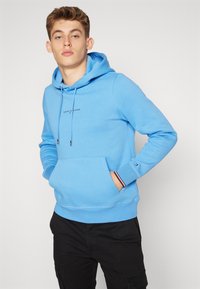 Felpa blu realizzata in morbido tessuto, con tasca frontale, cappuccio regolabile con coulisse e un piccolo logo sul petto. Pantaloni neri indossati sotto.