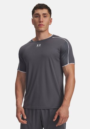 Mladý muž v priliehavej tmavosivej športovej tričku značky Under Armour s bielym lemovaním, stojaci pred jednoduchým svetlým pozadím.