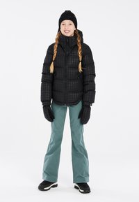 Giacca a vento nera con motivo a scacchi, abbinata a pantaloni da neve turchesi e guanti neri. Un caldo berretto completa l'abbigliamento invernale.