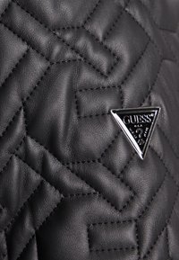 Cuero negro acolchado con un patrón de diamantes que presenta un logotipo triangular plateado de Guess. La textura es suave con acentos de costura visibles.