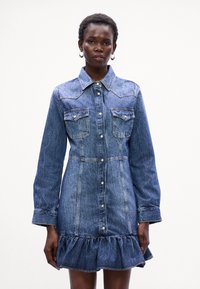 Vestido de ganga com corpete ajustado, mangas compridas, dois bolsos no peito e barra em camadas. Cor azul clara com detalhes de costura visíveis.