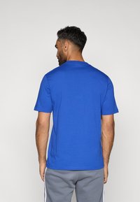 Βασιλικό μπλε κοντομάνικο μπλουζάκι από μαλακό ύφασμα, με σχέδιο λαιμόκοψης. Συνδυασμένο με γκρι φόρμες, προβάλλει μια χαλαρή στυλιστική επιλογή.