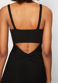 Femme portant une robe noire sans manches avec un détail torsadé et ouvert dans le dos, dévoilant le haut de son dos et ses épaules.