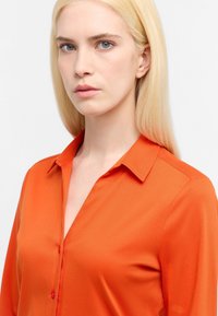 Camicia arancione con colletto, caratterizzata da una texture liscia e maniche lunghe. Dettagli notevoli includono i bottoni frontali dello stesso colore.